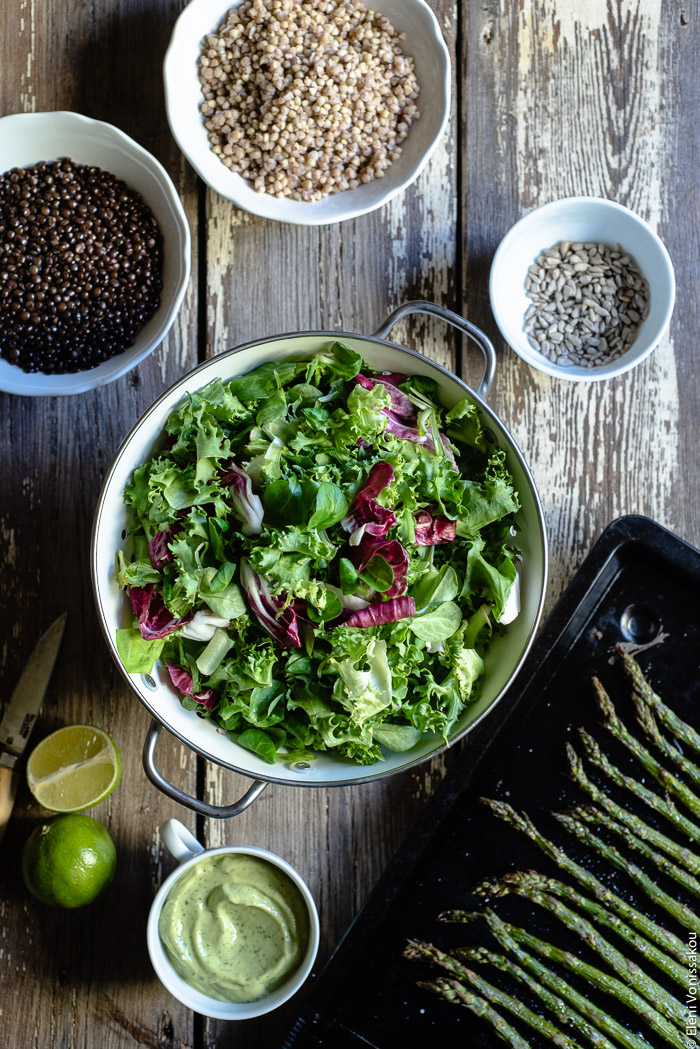 Green Salad with Buckwheat, Beluga Lentils, Roasted Asparagus and Avocado Dressing www.thefoodiecorner.gr Photo description: A top view of some ingredients on a wooden surface. To the top a bowl of cooked buckwheat, to the right a tiny bowl of sunflower seeds, to the bottom right a baking tray with roasted asparagus, to the bottom left a cup of creamy dressing, a knife and a lime, to the top left a bowl of cooked beluga lentils. In the centre a colander with mixed salad leaves.