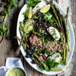 Green Salad with Buckwheat, Beluga Lentils, Roasted Asparagus and Avocado Dressing www.thefoodiecorner.gr Photo description: A platter on an old wooden surface, some parsley leaves and blueberries scattered beside it. To the bottom of the photo a cup with creamy dressing in it, sitting on a bunched up linen napkin. On the platter are green salad leaves and arranged on top of those are some buckwheat, beluga lentils, roasted asparagus, blueberries, slices of lime, blobs of fresh white cheese and a spoon with dressing nestled among them.