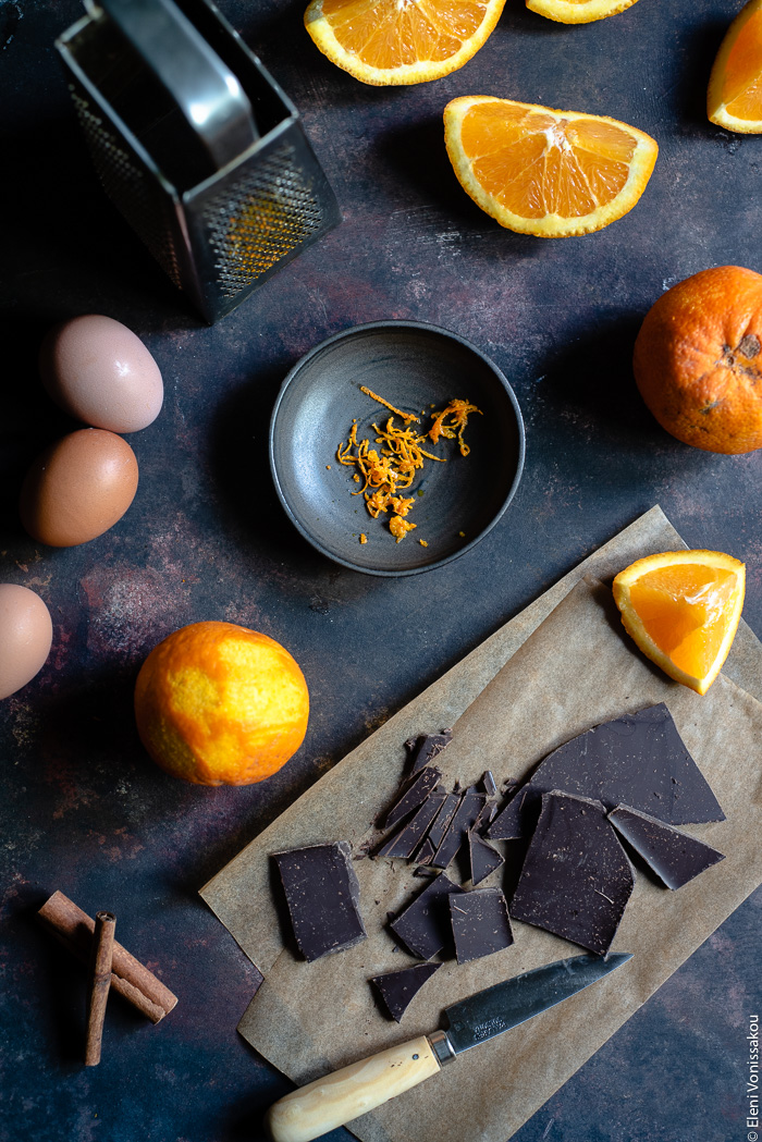 Chocolate Orange Curd – The Easy Way www.thefoodiecorner.gr Photo description: Top view of some ingredients spread out on a dark surface. Some pieces of orange in the top right corner, some pieces of chocolate and a knife on some grease-proof paper in the bottom right corner. At the bottom left are some cinnamon sticks , above those some eggs and a zested orange. In the top left corner a box grater and in the centre of the image is a small bowl with zest.