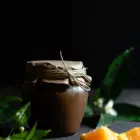 Chocolate Orange Curd – The Easy Way www.thefoodiecorner.gr Photo description: A jar of chocolate orange curd with a paper cover over the lid secured with string. In the foreground, some pieces of orange and some leaves from an orange tree. In the background, some more leaves with orange blossoms.