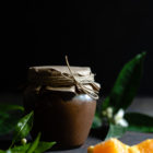 Εύκολη Κρέμα Σοκολάτα Πορτοκάλι www.thefoodiecorner.gr Photo description: A jar of chocolate orange curd with a paper cover over the lid secured with string. In the foreground, some pieces of orange and some leaves from an orange tree. In the background, some more leaves with orange blossoms.