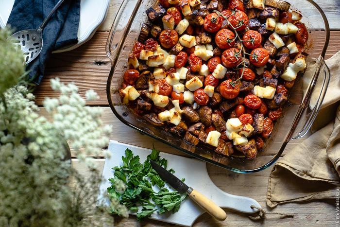 Roast Aubergine (Eggplant) and Cherry Tomato Pasta with Halloumi Cheese www.thefoodiecorner.gr Photo description: top view of vegetables (aubergine and cherry tomatoes) in a baking dish, roasted until soft. In one corner of the photo a bunch of Queen Anne’s Lace flowers. Beside the baking dish a small chopping board with a knife and some parsley.