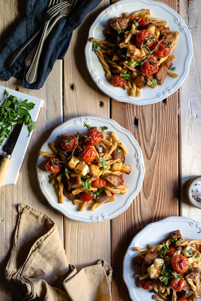 Roast Aubergine (Eggplant) and Cherry Tomato Pasta with Halloumi Cheese www.thefoodiecorner.gr Photo description: Top view of three plates of pasta on a wooden surface. In one corner of the image is a linen napkin and in another are some forks. On one side (barely visible) is a small chopping board with parsley and a knife. 