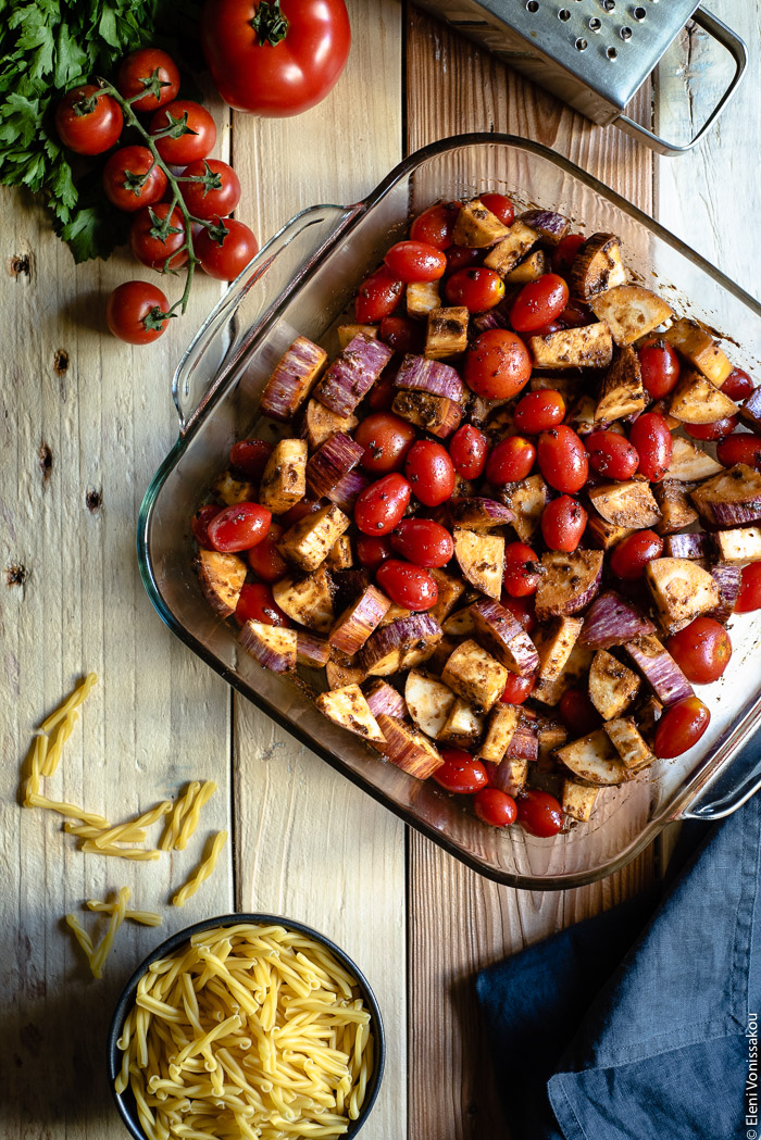 Roast Aubergine (Eggplant) and Cherry Tomato Pasta with Halloumi Cheese www.thefoodiecorner.gr Photo description: top view of raw vegetables in a baking dish. To the top of the photo some cherry tomatoes and a box grater (barely visible) and to the bottom a bunched up linen napkin and a small bowl of uncooked pasta. 