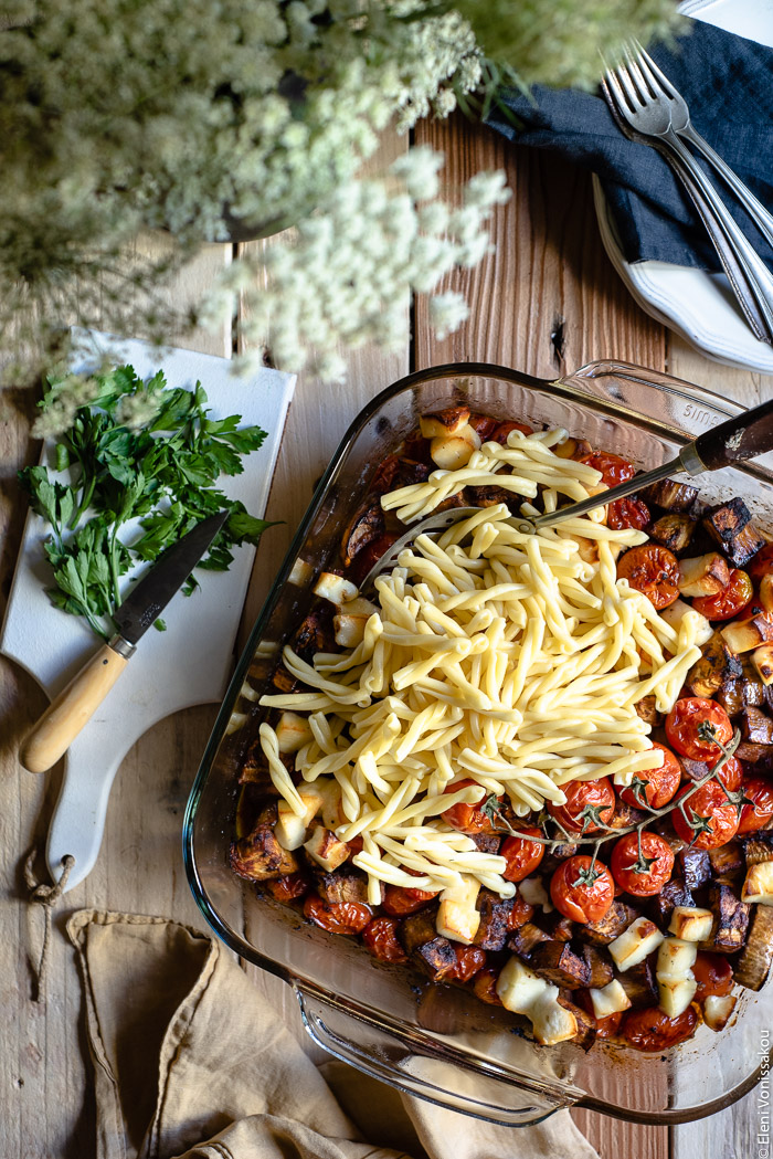 Roast Aubergine (Eggplant) and Cherry Tomato Pasta with Halloumi Cheese www.thefoodiecorner.gr Photo description: top view of vegetables (aubergine and cherry tomatoes) in a baking dish, roasted until soft. Boiled pasta has been added on top, in one corner of the dish, ready to be mixed in.