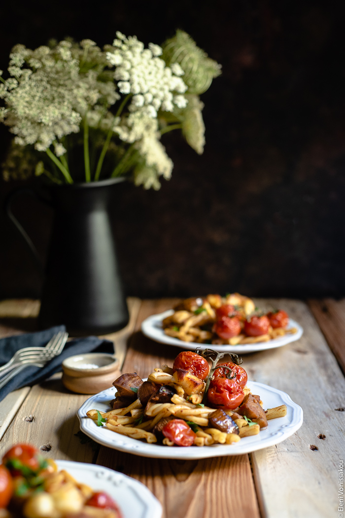 Roast Aubergine (Eggplant) and Cherry Tomato Pasta with Halloumi Cheese www.thefoodiecorner.gr Photo description: Three quarter view of plates filled with pasta, one of them with a small bunch of roasted cherry tomatoes on top. In the background a jug with Queen Anne’s Lace flowers.