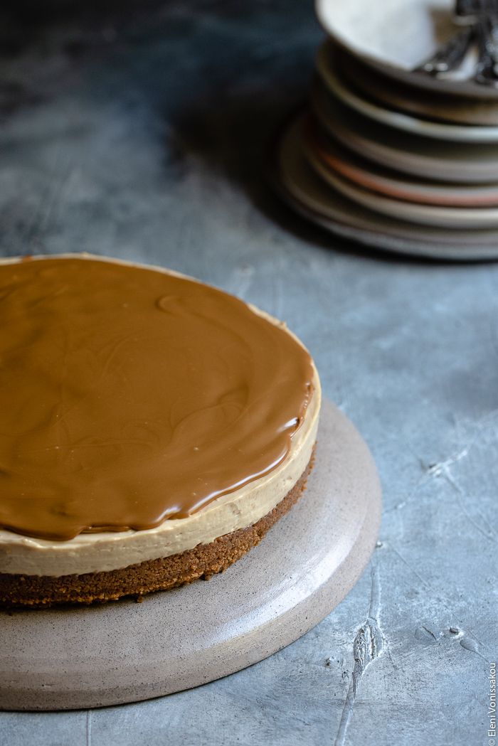 Εύκολο Τσίζκεϊκ με Άλειμμα Μπισκότου Speculoos (Χωρίς Ψήσιμο) www.thefoodiecorner.gr Photo description: A whole speculoos cheesecake with a layer of melted speculoos spread on top. In the background is a stack of ceramic plates.