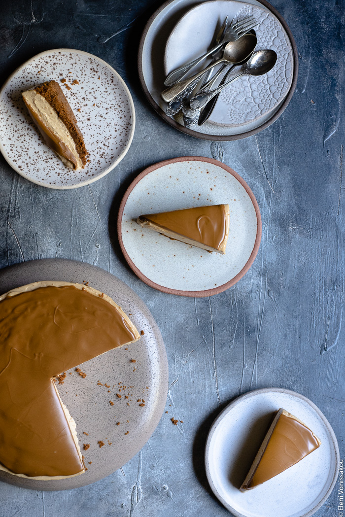 Easy Speculoos Cheesecake www.thefoodiecorner.gr Photo description: A cut speculoos cheesecake to the bottom left of the image and three plates surrounding it, each containing a slice of cheesecake. In the top right corner a couple of stacked plates with some spoons on the top one.