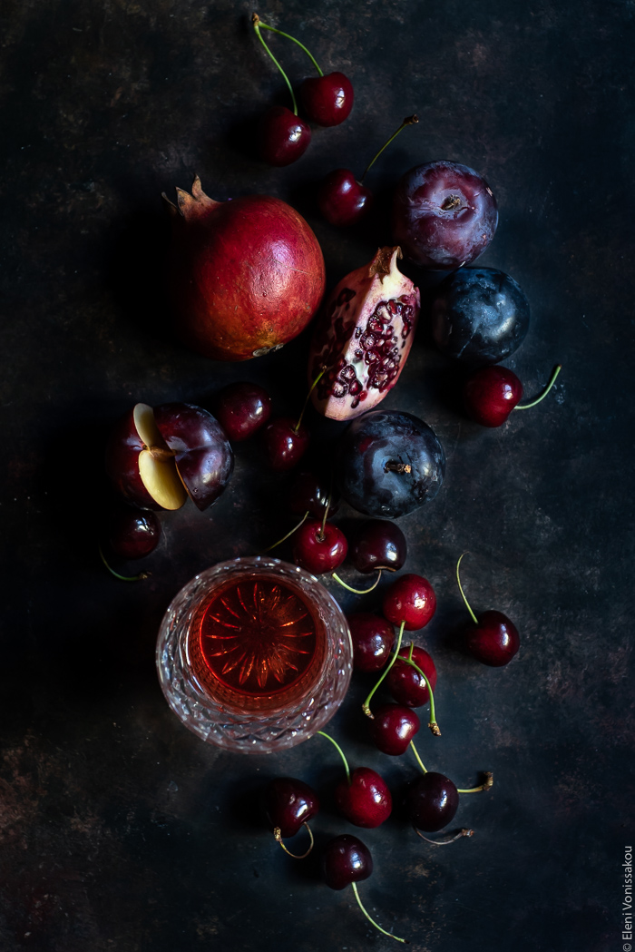 Pomegranate and Sour Cherry Rosé Sangria www.thefoodiecorner.gr Photo description: Overhead view of some pomegranate, cherries, plums and a glass of rose on a dark surface.