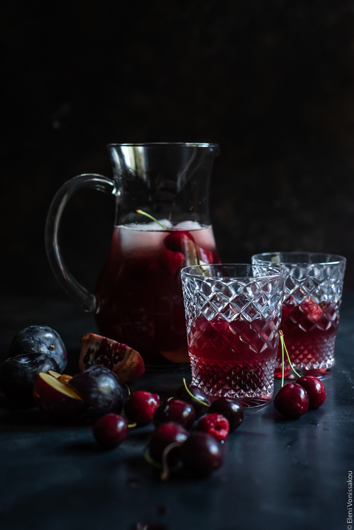 Pomegranate and Sour Cherry Rosé Sangria www.thefoodiecorner.gr Photo description: A jug of sangria in the background with two crystal glasses in front of it, each filled with the dark pink drink, and some red fruit on a dark surface.