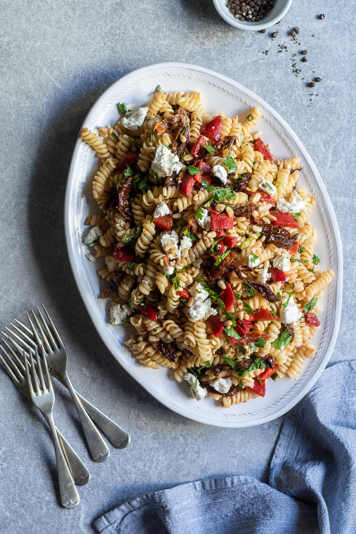 Pasta Salad with Sundried Tomatoes, Roasted Red Pepper, Feta and Tangy Goat’s Yoghurt Dressing www.thefoodiecorner.gr Photo description: A long platter with tossed pasta salad. To the bottom of the photo are a bunched up linen tea towel and some forks. 