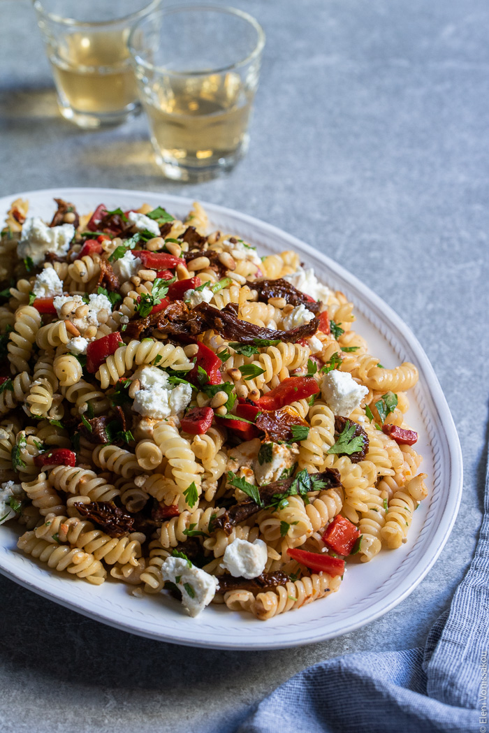 Pasta Salad with Sundried Tomatoes, Roasted Red Pepper, Feta and Tangy Goat’s Yoghurt Dressing www.thefoodiecorner.gr Photo description: A close view of a platter with tossed pasta salad. In the background two small glasses of white wine.