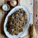 Plant-Based Slow Cooker Balsamic Mushroom Orzotto with Cashews and Sage www.thefoodiecorner.gr Photo description: A large platter with balsamic mushroom orzotto. To the bottom right is a bunched up linen napkin. To the top left are three small bowls, one wooden one with nutritional yeast, one ceramic with salt and one brass with dried sage.