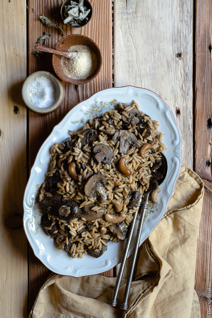 Plant-Based Slow Cooker Balsamic Mushroom Orzotto with Cashews and Sage www.thefoodiecorner.gr Photo description: A large platter of balsamic mushroom orzotto with two serving spoons lying at the side of the food. To the bottom right of the image is a bunched up linen napkin. To the top left are three small bowls, one wooden one with nutritional yeast, one ceramic with salt and one brass with dried sage.