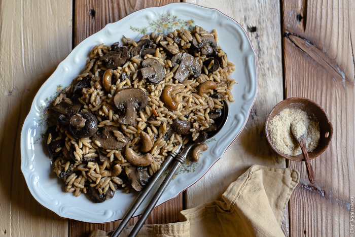 Plant-Based Slow Cooker Balsamic Mushroom Orzotto with Cashews and Sage www.thefoodiecorner.gr photo description: A large platter of balsamic mushroom orzotto, two serving spoons placed to the side of the food. To the bottom right of the image is a bunched up linen napkin and a small wooden bowl with nutritional yeast flakes.