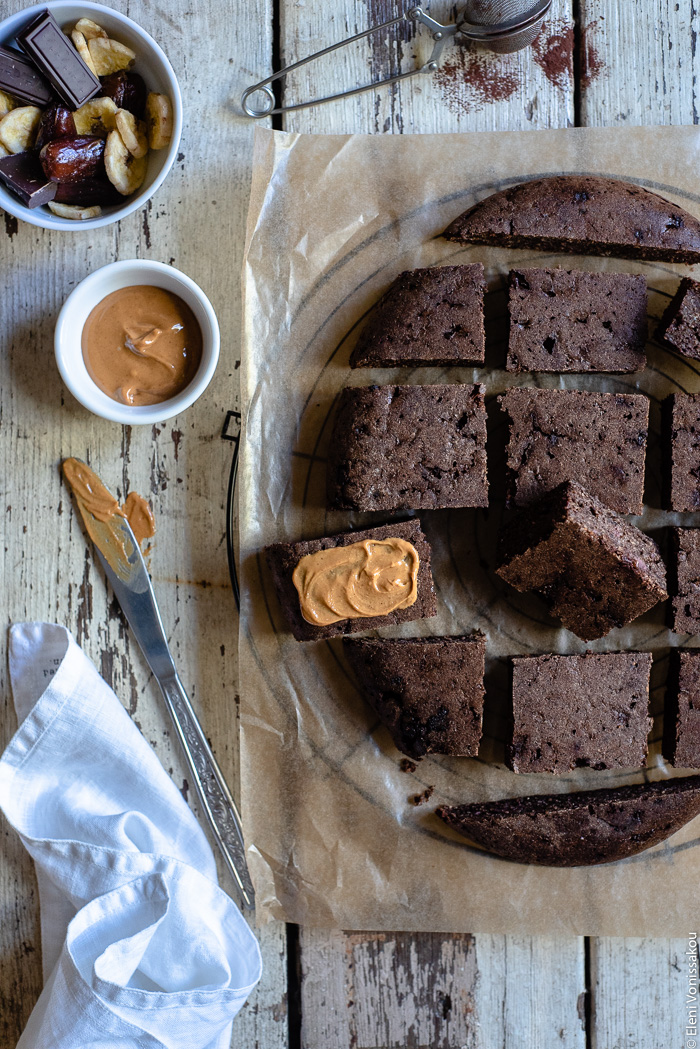 Plant-Based, Date-Sweetened Chocolate Banana Oat Cake-Bars, in the Slow Cooker www.thefoodiecorner.gr Photo description: Chocolate Banana Oat Cake-Bars cut up and arranged (in the shape of the slow cooker) on a piece of grease-proof paper on a wire rack. To the top left of the photo is a bowl of dates, chocolate and banana chips, further down is a small bowl of peanut butter and a knife laying on the wooden surface. The knife is smeared with peanut butter. To the bottom left is a bunched up linen napkin. Some peanut butter has also been spread on one of the cake-bars.