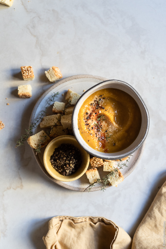 Slow Cooker Butternut Squash and Red Lentil Soup - Low Waste and Minimal Prep www.thefoodiecorner.gr Photo description: A bowl of soup sitting on a small plate along with a tiny bowl of pepper and some croutons. The soup is garnished with black pepper, paprika and nutritional yeast. To the bottom of the image is a bunched up linen tea towel. 