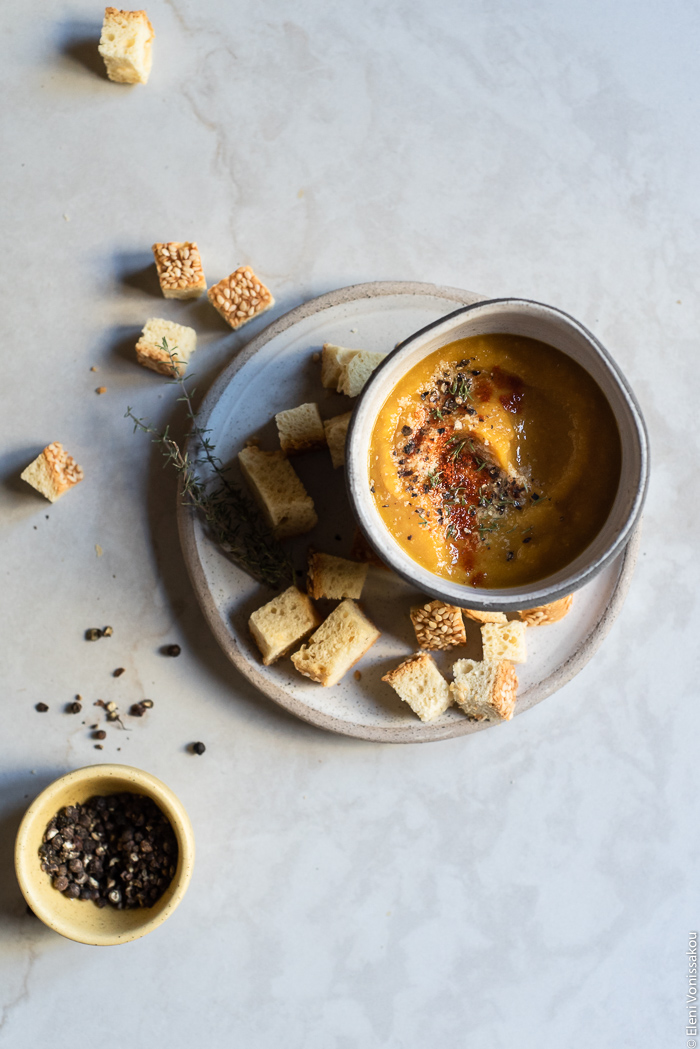 Slow Cooker Butternut Squash and Red Lentil Soup - Low Waste and Minimal Prep www.thefoodiecorner.gr Photo description: A bowl of soup sitting on a small plate along with some croutons. The soup is garnished with black pepper, paprika and nutritional yeast. To the bottom left of the image is a tiny bowl of black pepper. 