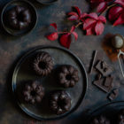 Chocolate and Beetroot Mini Bundt Cakes with a Quick Ganache (Plant Based) www.thefoodiecorner.gr Photo description: Overhead view of mini bundt cakes on a dark plate. Around the plate are some pretty autumn leaves, some chopped chocolate and some cocoa powder. In the two corners of the photo are some more bundt cakes.