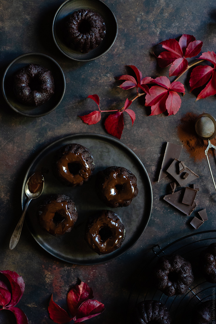 Chocolate and Beetroot Mini Bundt Cakes with a Quick Ganache (Plant Based) www.thefoodiecorner.gr Photo description: Overhead view of mini bundt cakes with a ganache topping on a dark plate. Around the plate are some pretty autumn leaves, some chopped chocolate and some cocoa powder. In the two corners of the photo are some more bundt cakes.