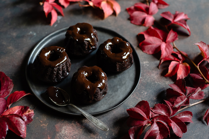 Chocolate and Beetroot Mini Bundt Cakes with a Quick Ganache (Plant Based) www.thefoodiecorner.gr Photo description: A ¾ view of a plate with 4 mini bundt cakes on it. The cakes have been topped with ganache and the spoon used is lying on the plate next to them.