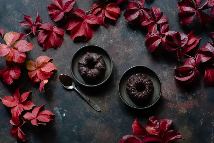 Chocolate and Beetroot Mini Bundt Cakes with a Quick Ganache (Plant Based) www.thefoodiecorner.gr Photo description: An overhead view of two mini bundt cakes each on a small ceramic cake. To the side of one of the plates is a spoon with some ganache on it. Scattered all around are some pretty autumn leaves.
