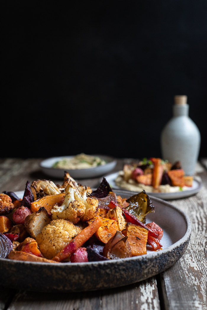 Cumin Roasted Root Vegetable Medley. My Winter “Briam”. www.thefoodiecorner.gr Photo description: A side view of a ceramic dish full of roasted root vegetables against a dark background. Behind the dish are some smaller plates of food and a ceramic bottle.