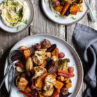 Cumin Roasted Root Vegetable Medley. My Winter “Briam”. www.thefoodiecorner.gr Photo description: A large ceramic dish full of roasted root vegetables on an worn wooden surface. To the top of the dish are two smaller ceramic plates, one with hummus the other with a portion of veg. A linen napkin lies on the right side of the large dish.