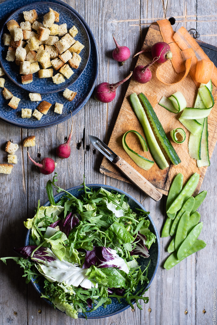 Green Salad with Cucumber, Radishes, Homemade Croutons and Grape Must Vinaigrette www.thefoodiecorner.gr Photo description: A bowl of salad leaves next to a chopping board with a peeler and slices of cucumber and carrot on it. To the top of the photo is a small plate with croutons.