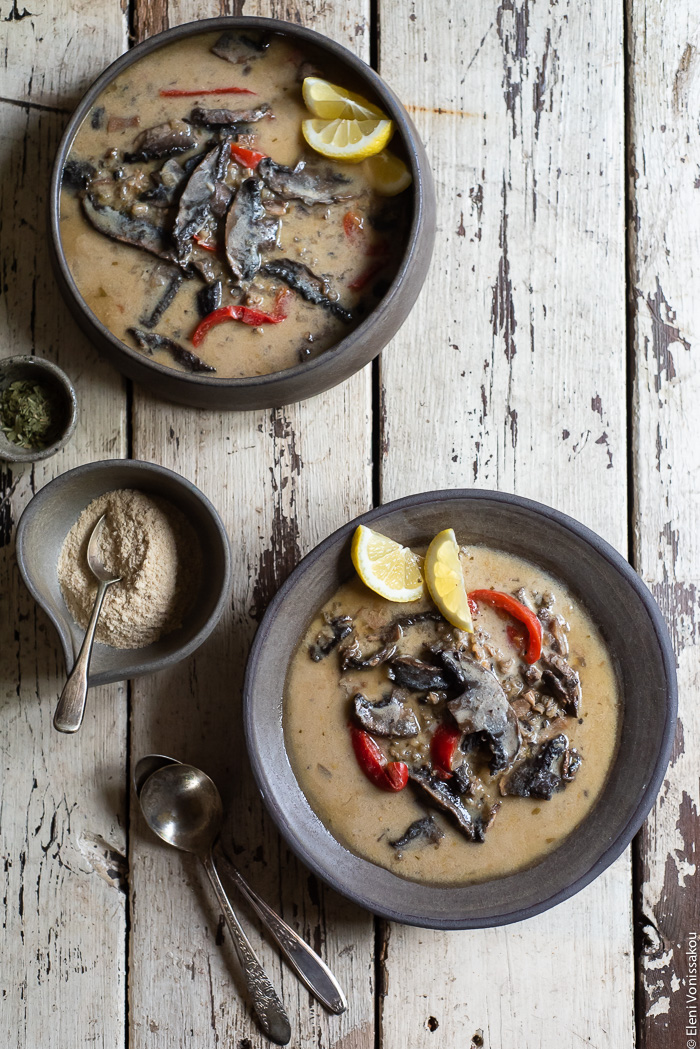 Plant-Based Creamy Mushroom and Wild Rice Soup in the Slow Cooker www.thefoodiecorner.gr Photo description: Two bowls of creamy mushroom and wild rice soup with red peppers, garnished with some slices of lemon. To the left a bowl with nutritional yeast flakes and a couple of vintage spoons. Everything is placed on a distressed wooden surface. 