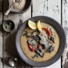 Plant-Based Creamy Mushroom and Wild Rice Soup in the Slow Cooker www.thefoodiecorner.gr Photo description: A ceramic bowl of creamy mushroom and wild rice soup with red peppers. Two slices of lemon sit at the edge of the bowl. To the left a small bowl with nutritional yeast flakes and a tiny bowl of dried tarragon. To the bottom of the main dish are two vintage spoons. Everything is placed on a distressed wooden surface.