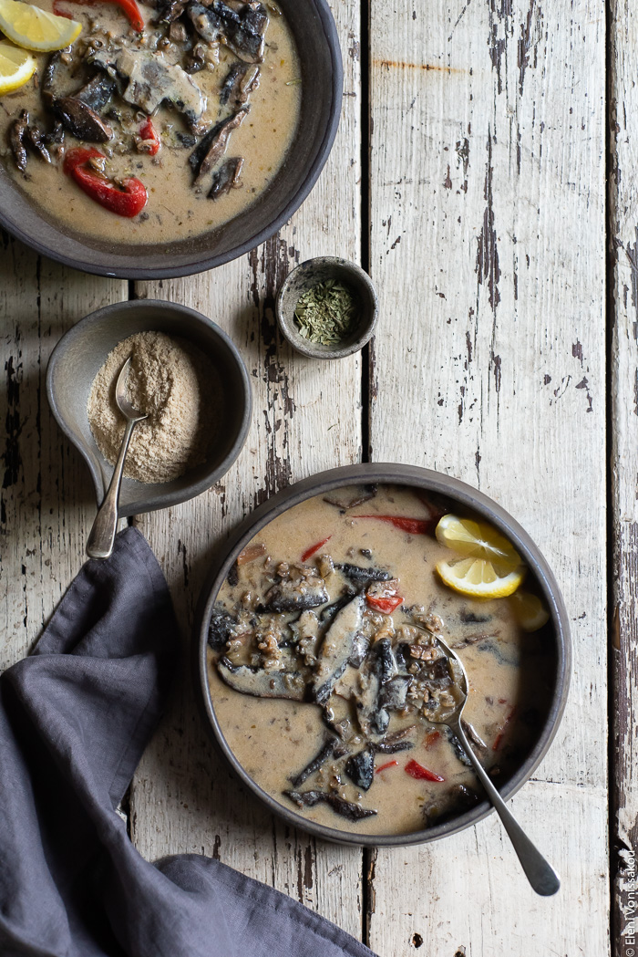 Plant-Based Creamy Mushroom and Wild Rice Soup in the Slow Cooker www.thefoodiecorner.gr Photo description: Two bowls of creamy mushroom and wild rice soup with red peppers, garnished with some slices of lemon. One bowl is barely visible in the corner of the image, the other has a spoon inside the soup. To the left is a small bowl with nutritional yeast, a tiny bowl with dried tarragon and a bunched up linen napkin.