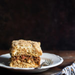 Pastitsio (Greek Pasta Bake) with Lentil and Mushroom Sauce, and Oat Milk Bechamel www.thefoodiecorner.gr Photo description: A side view of a plate with a slice of pastitsio on it, the layers of pasta and lentil sauce clearly visible. To the right, a bunched up tea towel.