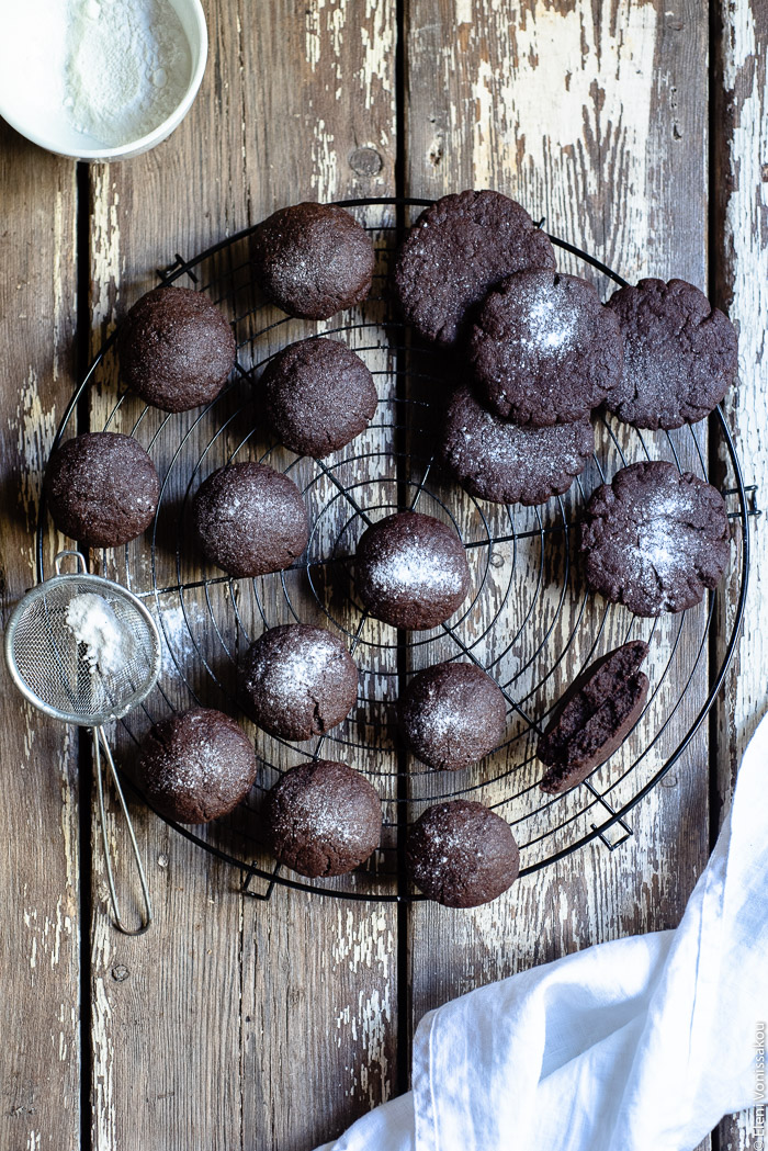 Chocolate Tahini Olive Oil Cookies or Brownies (One Recipe, Two Treats) www.thefoodiecorner.gr Photo description: A top view of a cooking rack full of chocolate biscuits/cookies. Some of them are dome shaped and some are round and flat. Some of them have been sprinkled with icing sugar. To the left is a small strainer with sugar in it and to the top left is a small bowl with extra icing sugar. 