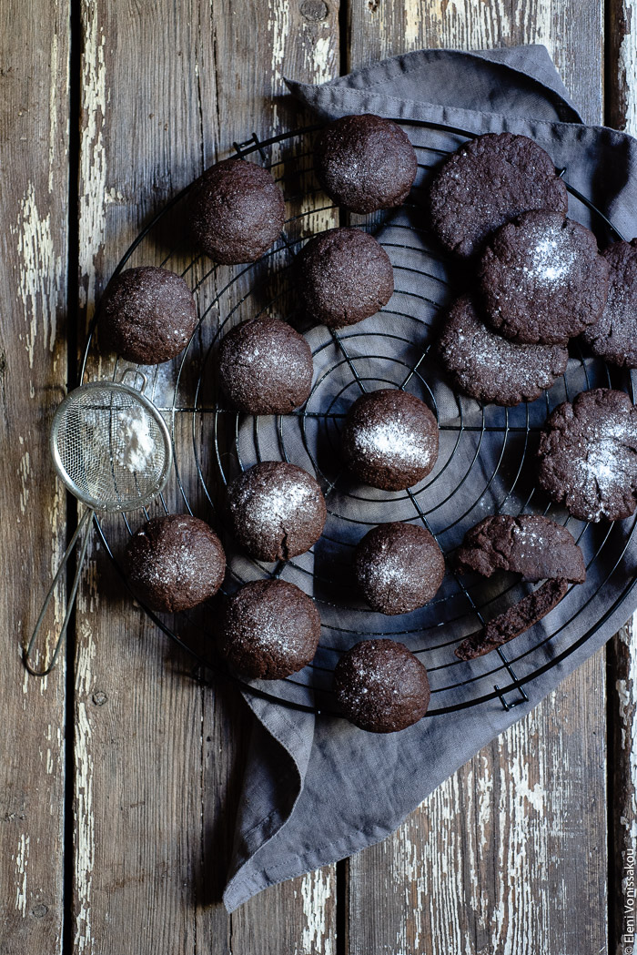 Chocolate Tahini Olive Oil Cookies or Brownies (One Recipe, Two Treats) www.thefoodiecorner.gr Photo description: A top view of a cooking rack full of chocolate biscuits/cookies. Some of them are dome shaped and some are round and flat. Some of them have been sprinkled with icing sugar. To the left is a small strainer with sugar in it and beneath the rack is a bunched up linen napkin.