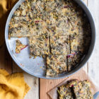 Easy Greek “Flour Pie” with Cornmeal, Spelt and Mixed Greens (Spinach and Beets) www.thefoodiecorner.gr Photo description: A large baking tin with a thin flour-pie inside, some pieces of it missing. To the bottom right of the tin is a piece of grease-proof paper with a couple of slices of the pie and a knife on it. To the left of the tin are two linen napkins. Everything is on a surface made of wooden planks.