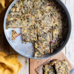 Easy Greek “Flour Pie” with Cornmeal, Spelt and Mixed Greens (Spinach and Beets) www.thefoodiecorner.gr Photo description: A large baking tin with a thin flour-pie inside, some pieces of it missing. To the bottom right of the tin is a piece of grease-proof paper with a couple of slices of the pie and a knife on it. To the left of the tin are two linen napkins. Everything is on a surface made of wooden planks.