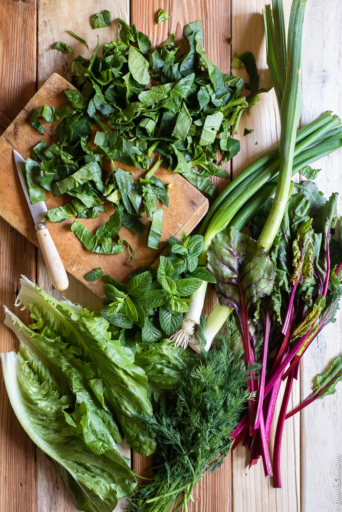 Easy Greek “Flour Pie” with Cornmeal, Spelt and Mixed Greens (Spinach and Beets) www.thefoodiecorner.gr Photo description: Lettuce leaves, beet leaves with their stalks, dill, spring onions and leeks all layed out on a wooden surface. Just above them is a chopping board with some chopped spinach and a knife on it.