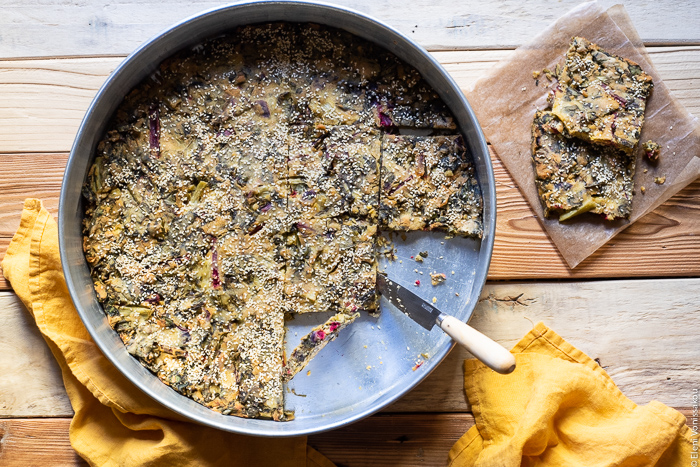 Easy Greek “Flour Pie” with Cornmeal, Spelt and Mixed Greens (Spinach and Beets) www.thefoodiecorner.gr Photo description: A large baking tin containing a cooked greens pie. Some slices have been removed and placed on a piece of baking paper at the side of the tin. A knife is lying over the edge of the tin, its tip touching the pie. To the bottom of the tin are two linen napkins.