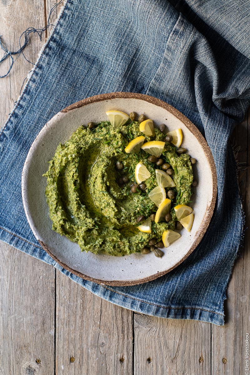 Greek Island Garlicky Parsley Dip with Easy Pitta Chips www.thefoodiecorner.gr Photo description: A bowl of parsley dip with a garnish of lemon, capers and chives, sitting on a piece of denim cloth.