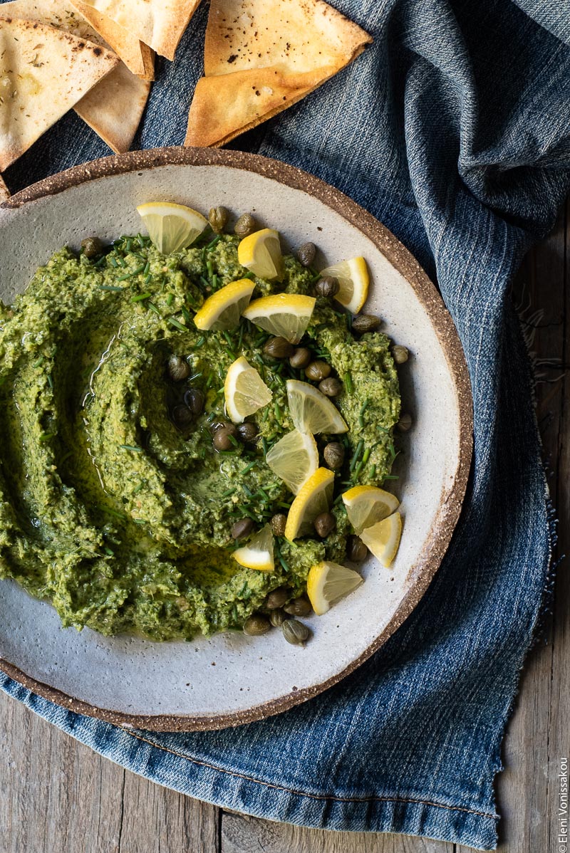 Greek Island Garlicky Parsley Dip with Easy Pitta Chips www.thefoodiecorner.gr Photo description: A close view of a bowl of parsley dip garnished with lemon pieces, capers and chopped chives. To the top of the bowl are some pitta chips. Everything is sitting on a piece of denim cloth.