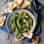 Greek Island Garlicky Parsley Dip with Easy Pitta Chips www.thefoodiecorner.gr Photo description: A bowl of parsley dip with a garnish of lemon, capers and chives. Surrounding the bowl are some triangular shaped pitta chips. The bowl and chips are sitting on a piece of denim cloth. At the side of the bowl are a small spoon with some dip on it and a chip with dip slathered on top.