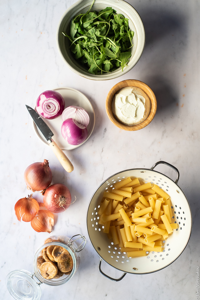 Caramelised Onion, Fig and Fresh Cheese Pasta with Arugula www.thefoodiecorner.gr Photo description: A bowl of arugula (rocket), a small plate with two peeled onions and a knife on it, a small bowl of fresh cheese, two whole onions and some onions skins, a jar of dried figs and a colander of dried pasta. All are arranged on a marble surface.