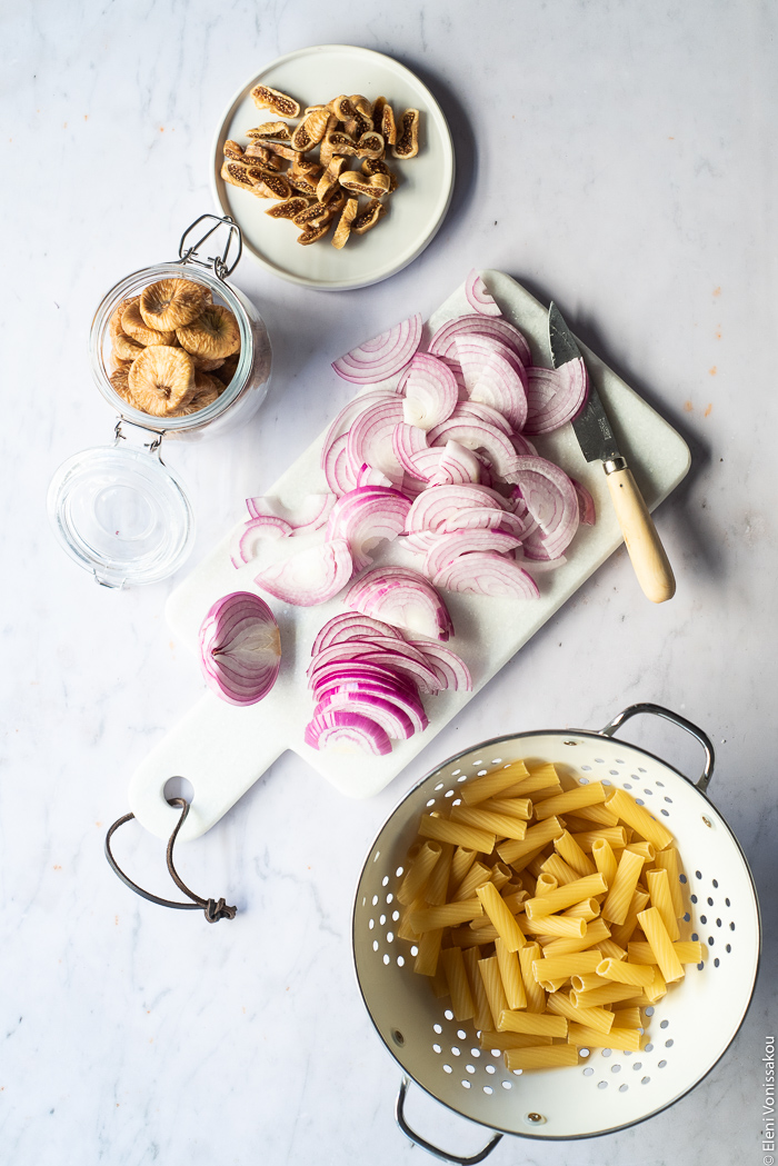 Caramelised Onion, Fig and Fresh Cheese Pasta with Arugula www.thefoodiecorner.gr Photo description: A small plate of chopped figs, a jar of whole dried figs, a chopping board with sliced onions and a knife on it, and a colander with dried rigatoni pasta inside.