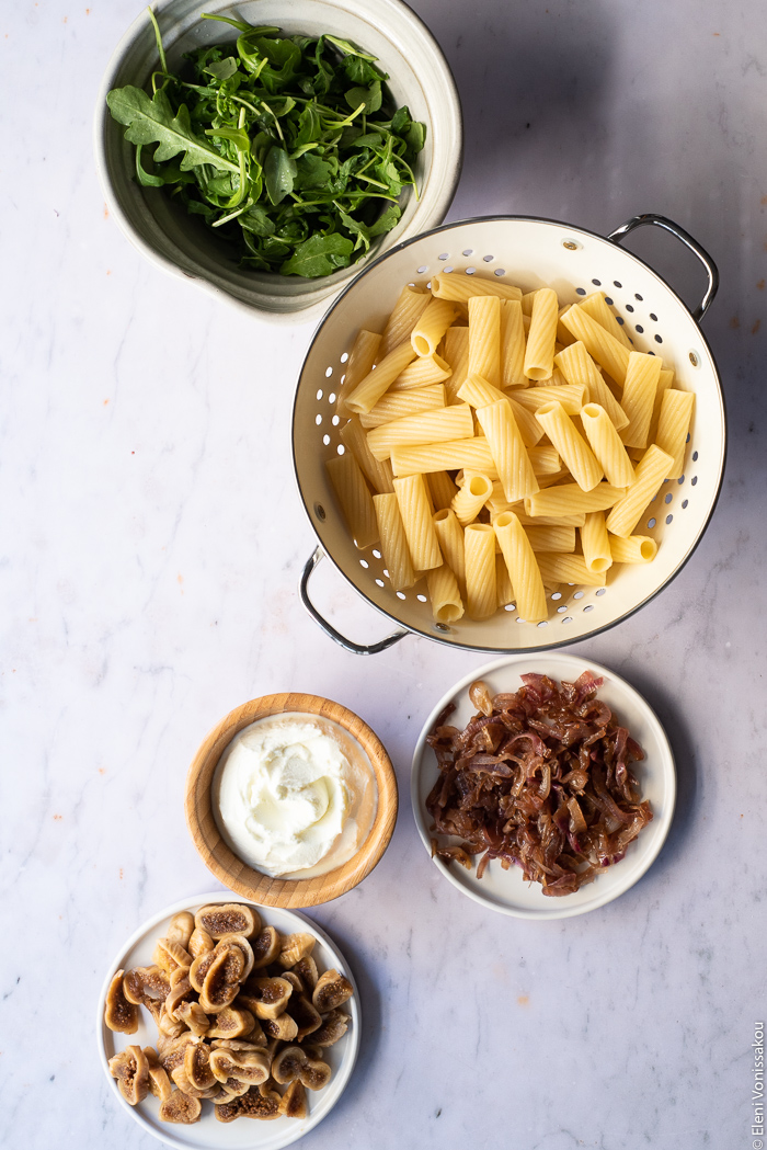 Caramelised Onion, Fig and Fresh Cheese Pasta with Arugula www.thefoodiecorner.gr Photo description: A bowl of arugula, a colander with boiled rigatoni pasta inside, a small plate of caramelised onions, a bowl of fresh cheese and a small plate of reconstituted figs.