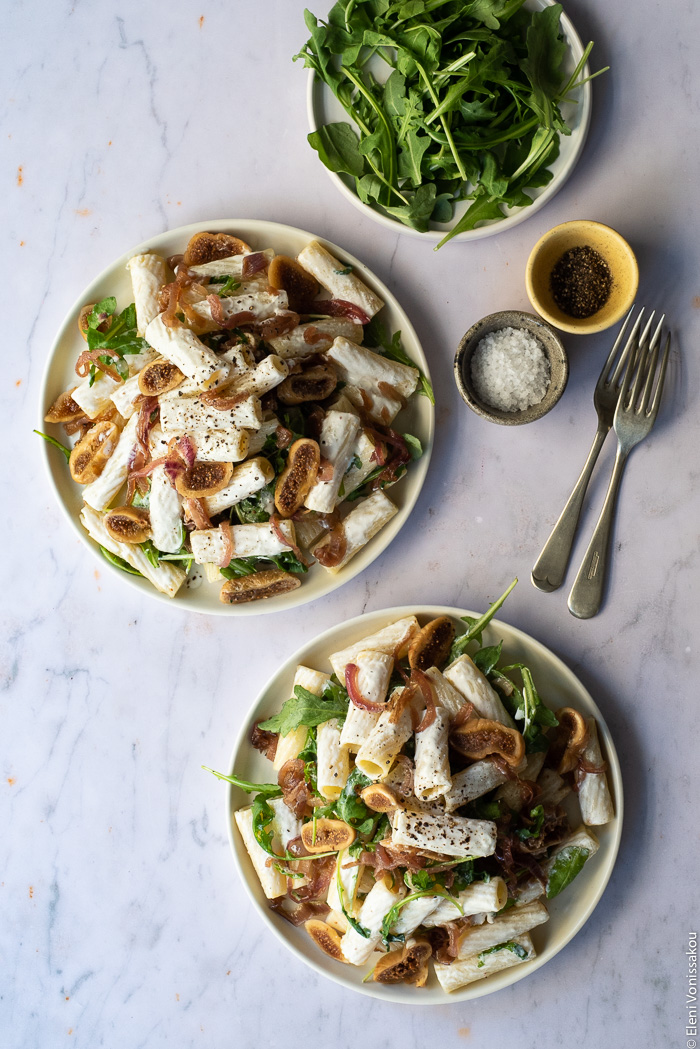 Caramelised Onion, Fig and Fresh Cheese Pasta with Arugula www.thefoodiecorner.gr Photo description: two plates of pasta on a marble surface. To the top right are two small bowls of salt and pepper, two forks and a small plate of extra arugula.