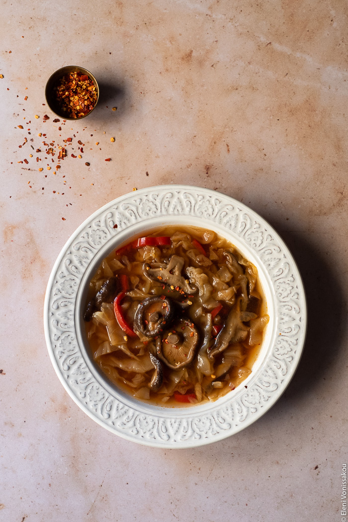 Slow Cooker Hot and Sour Cabbage Soup with Shiitake Mushrooms www.thefoodiecorner.gr Photo description: One bowl of soup on a marble surface. A bit further away is a tiny bowl of chilli flakes, with some of the flakes scattered on the marble.