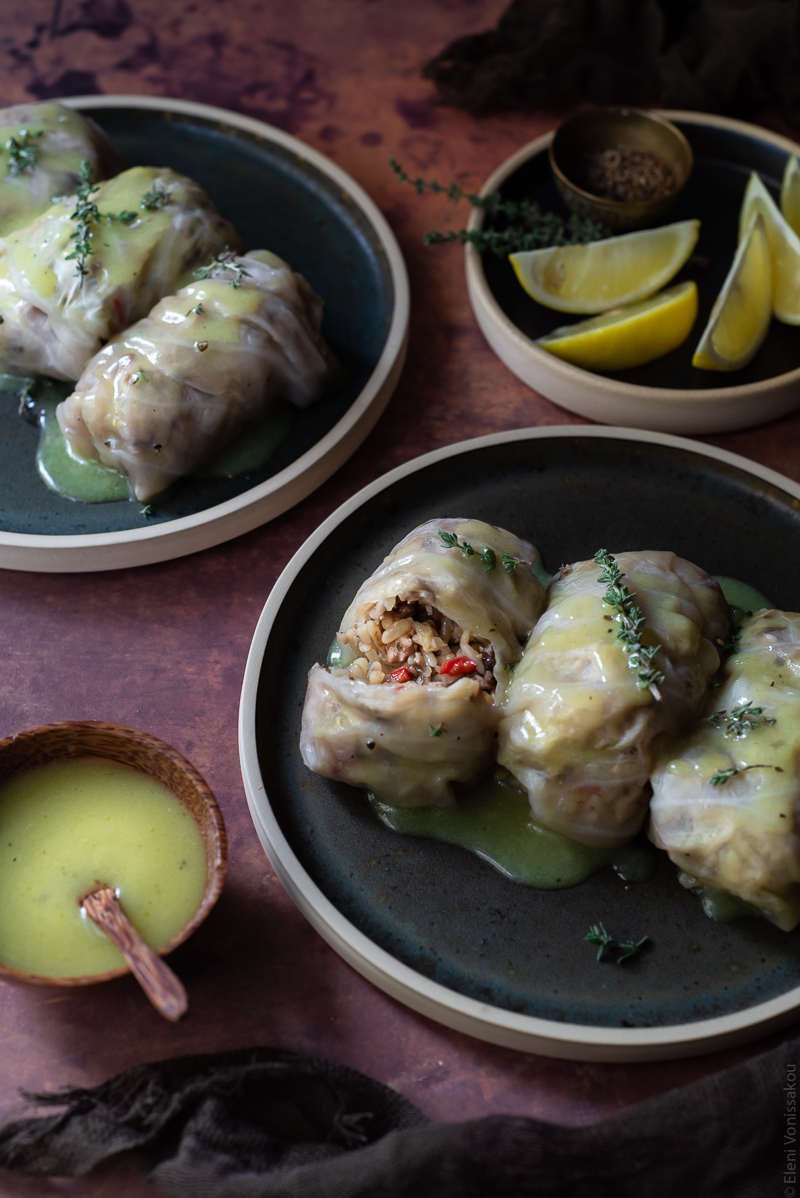 Shiitake, Rice and Truffle Oil Stuffed Cabbage Rolls with Lemon Vinaigrette www.thefoodiecorner.gr Photo description: A ¾ view of two plates with stuffed cabbage rolls on a rusty coloured surface. One of the rolls is cut showing the rice stuffing inside. In the top right corner of the image is a small plate with slices of lemon and a small bowl of vinaigrette.