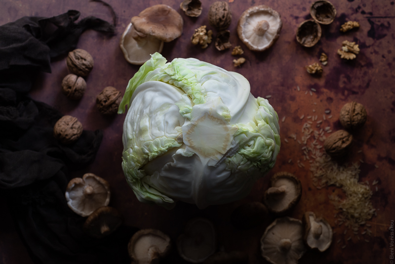 Shiitake, Rice and Truffle Oil Stuffed Cabbage Rolls with Lemon Vinaigrette www.thefoodiecorner.gr Photo description: A large cabbage sitting upside down (with the core looking up) in the centre of the image. Around it are some mushrooms and walnuts, all arranged on a rusty coloured surface.