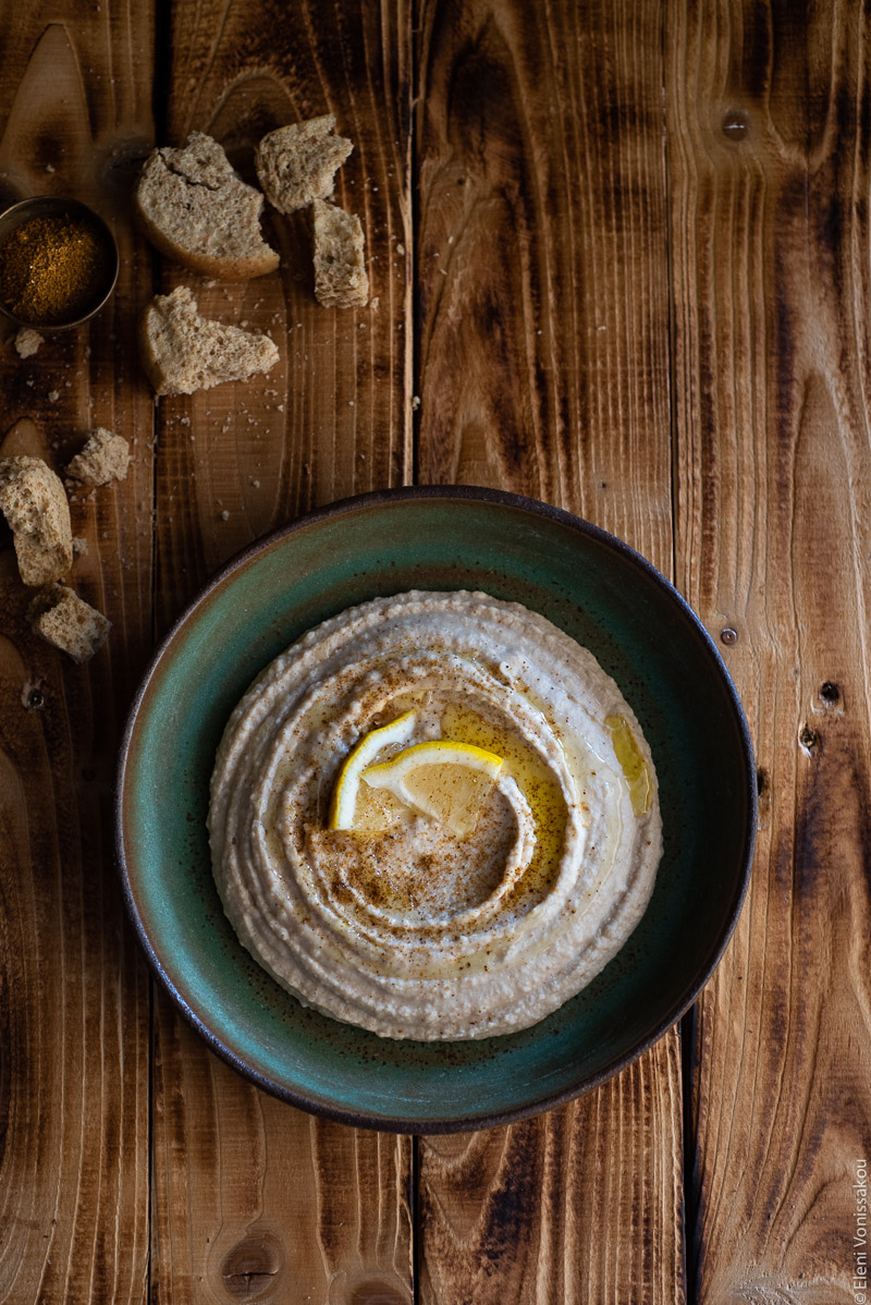 Hummus-Style White Bean Dip with Ras El Hanout www.thefoodiecorner.gr Photo description: A ceramic green dish with humms on a distressed wooden background. The hummus is dusted with ras el hanout, drizzled with olive oil and garnished with a couple of lemon wedges. To the top of the image directly on the wood are some more broken rusks and a tiny bowl of ras el hanout spice.